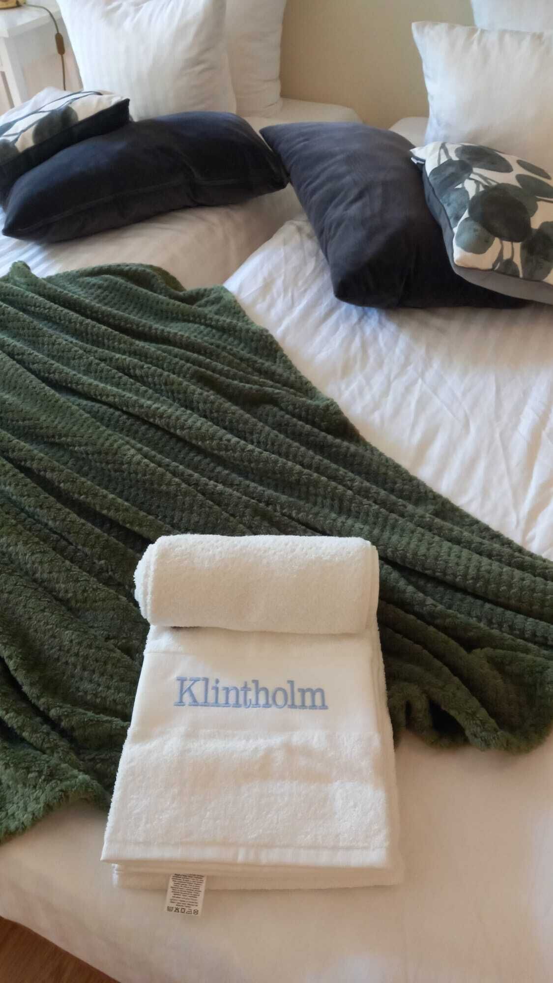 En pænt redt seng med et grønt tæppe, et hvidt sammenfoldet håndklæde med 'Klintholm' broderet på, og en pude med et blad-mønster.