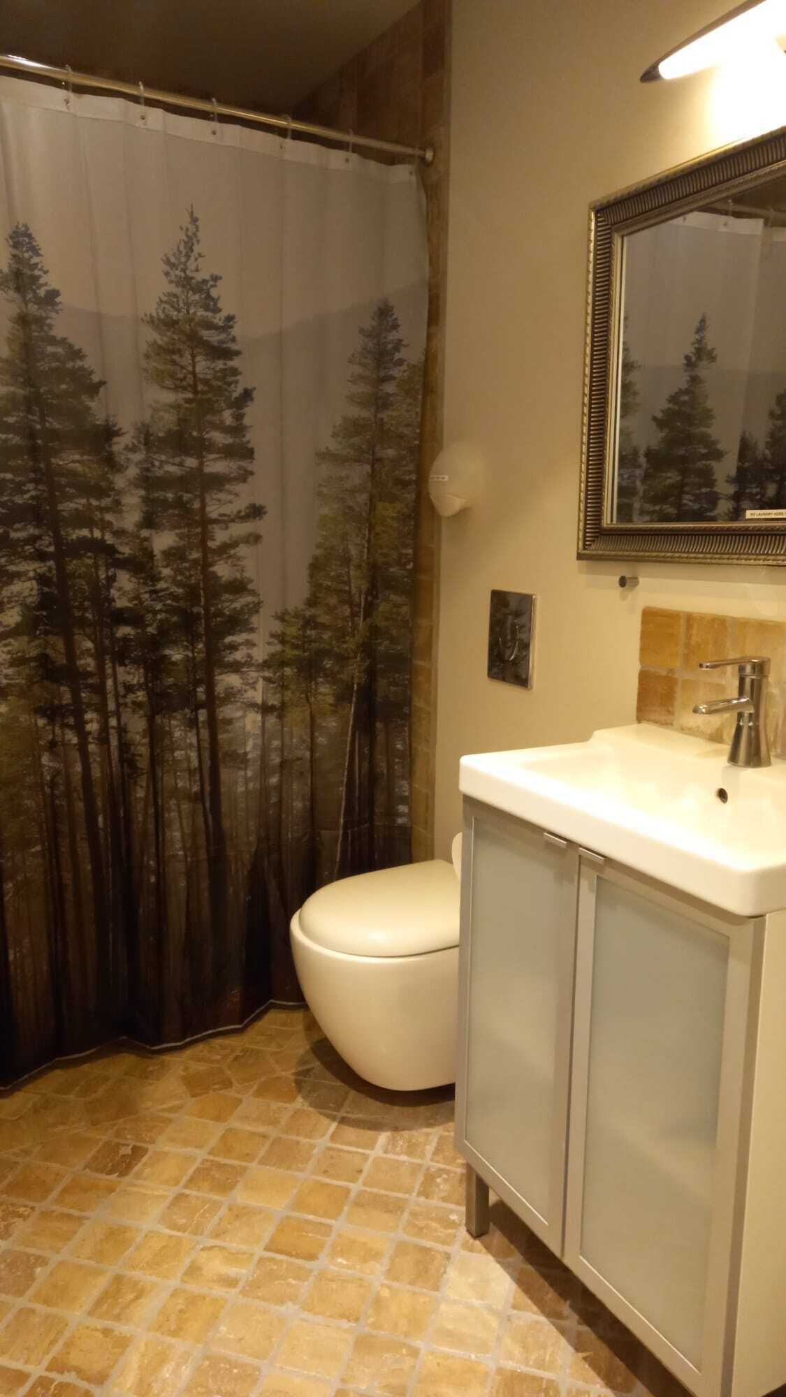 A bathroom with a shower curtain depicting trees, a toilet, a sink with a mirror, and a tiled floor.