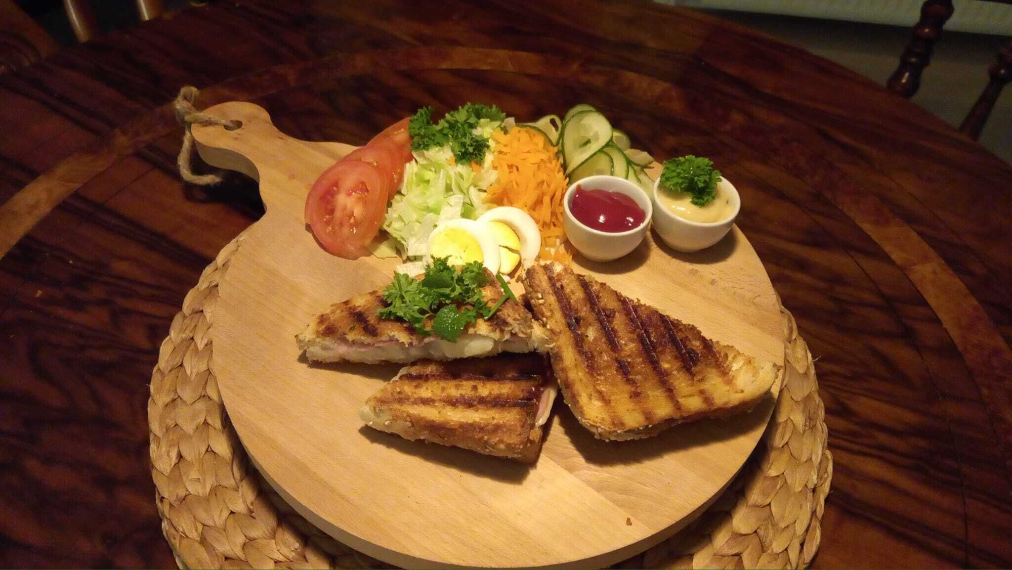 Una tabla de madera con sándwiches a la parrilla, una ensalada y dos recipientes pequeños con salsa.
