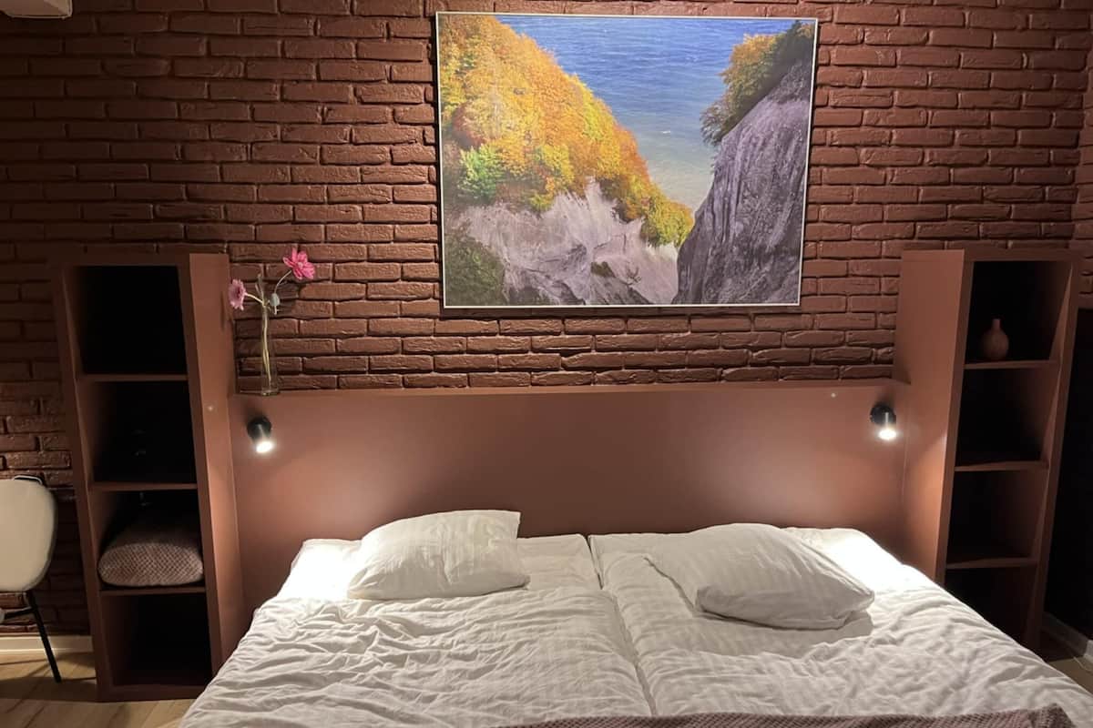 Ein Schlafzimmer mit Backsteinwand, einem Gemälde einer Küstenlandschaft, einem Bett mit weißer Bettwäsche und zwei Nachttischlampen.