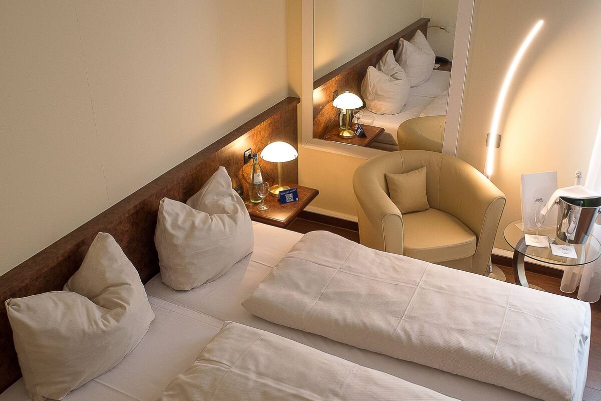 A hotel room with a bed, a chair, a nightstand with a lamp, and a mirror reflecting the bed and chair.
