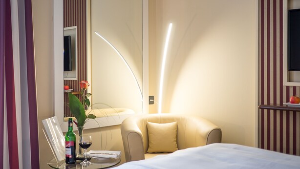 A hotel room with a bed, a round table with a bottle and glass, a chair, a sofa, a mirror, and a wall-mounted TV. - Hotel Primula (Troisdorf)