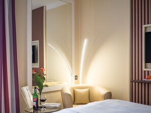 A hotel room with a bed, a round table with a bottle and glass, a chair, a sofa, a mirror, and a wall-mounted TV. - Hotel Primula (Troisdorf)