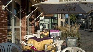 An outdoor seating area with tables and chairs, a large umbrella, and a sign for a restaurant. - Hotel Primula (Troisdorf)