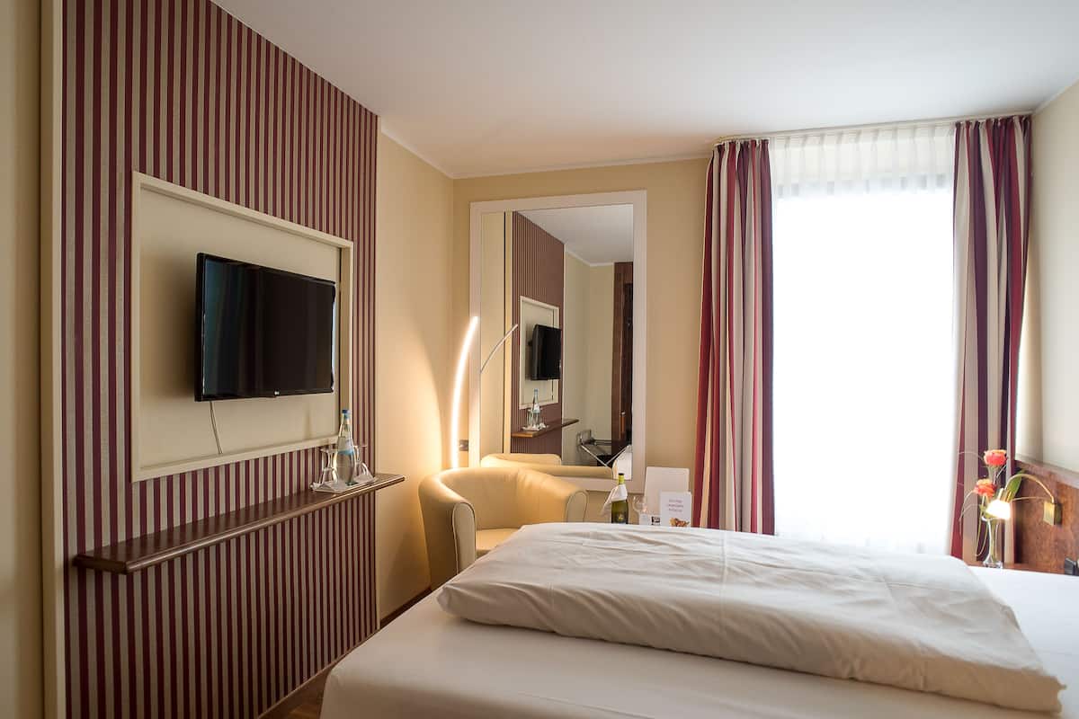 A hotel room with a bed, a TV mounted on the wall, a bedside table with a lamp, and a window with curtains.