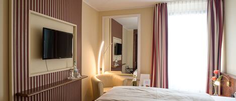 A hotel room with a bed, a TV mounted on the wall, a bedside table with a lamp, and a window with curtains.
