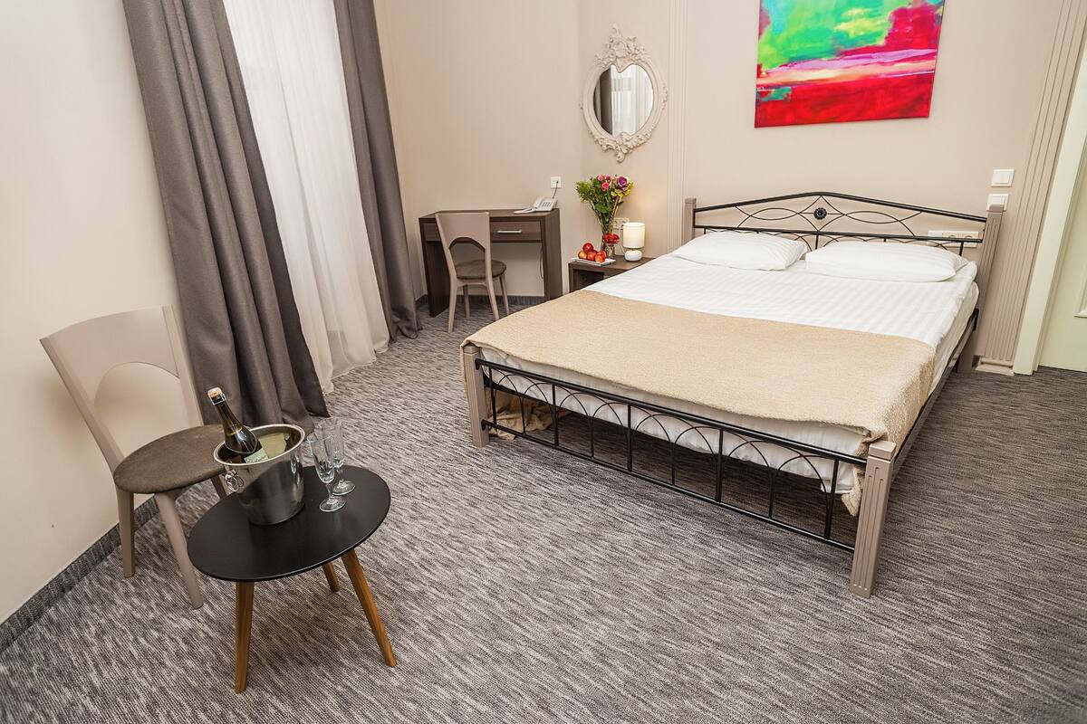 A hotel room with a bed, a desk, a chair, a small table with a bottle of champagne, and a colorful painting on the wall.