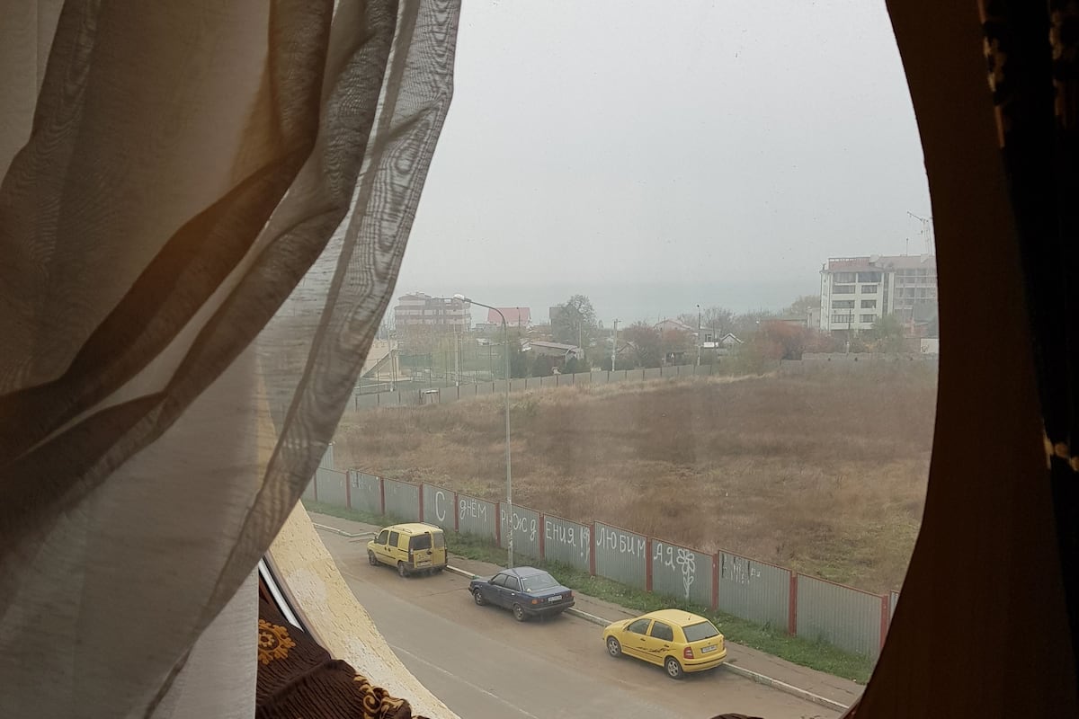 A view through a window with curtains, featuring a road with three vehicles and a fenced area with buildings in the background.