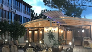 Un área de estar al aire libre con sillas y mesas de plástico, un porche cubierto y un edificio con grandes ventanales y unidades de aire acondicionado.