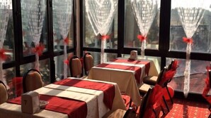 Un comedor con mesas dispuestas para cuatro, ventanas grandes con cortinas y un suelo cubierto de alfombra roja.