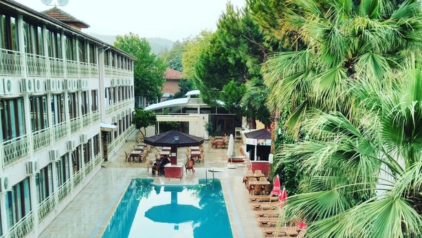 Un área de piscina con sillas de descanso y sombrillas, rodeada de follaje y un edificio con múltiples ventanas.