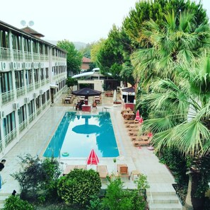 A pool area with lounge chairs and umbrellas, surrounded by greenery and a building with multiple windows. - OSKAR HOTEL (Denizli)