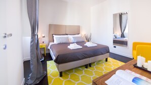 A hotel room with a bed, a bedside table with a lamp, a window with curtains, and a yellow chair. - Rent In Rome - Flo's Apartment (Rome)