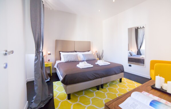 A hotel room with a bed, a bedside table with a lamp, a window with curtains, and a yellow chair. - Rent In Rome - Flo's Apartment (Rome)