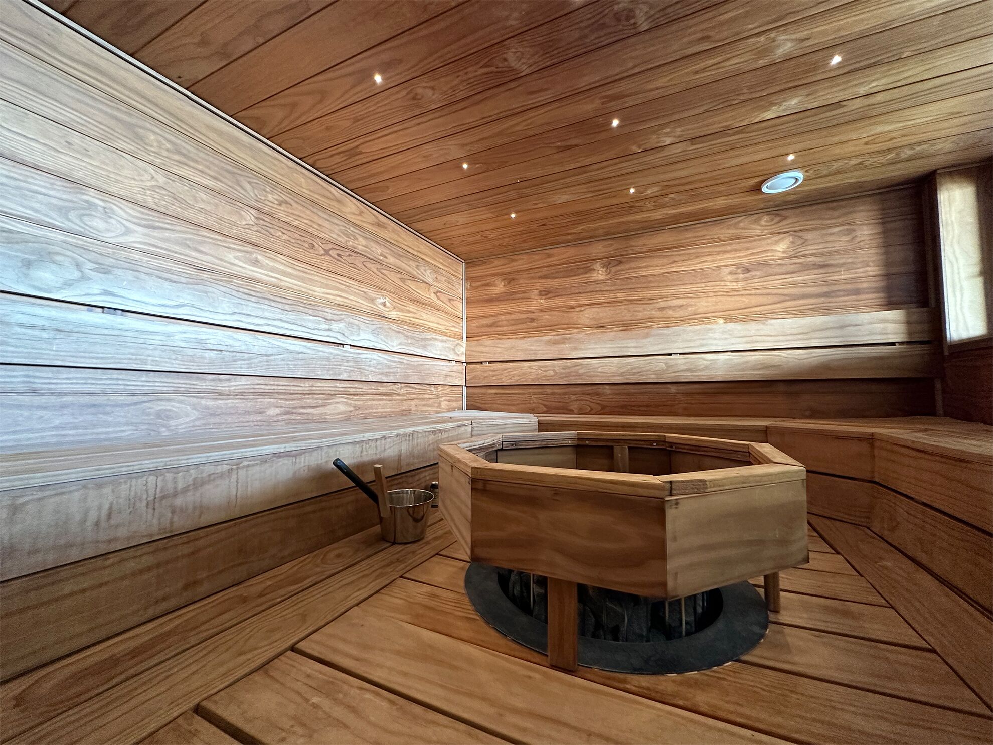 En sauna med trævægge og bænke, en central trækonstruktion og en lille metalspand.