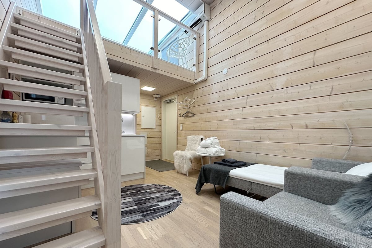 Un interior moderno con piso de madera, una escalera, un sofá, una mesita y vista al cielo a través de un tragaluz.