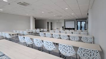 Sala de conferências com mesas longas e cadeiras brancas, um projetor e um quadro branco.