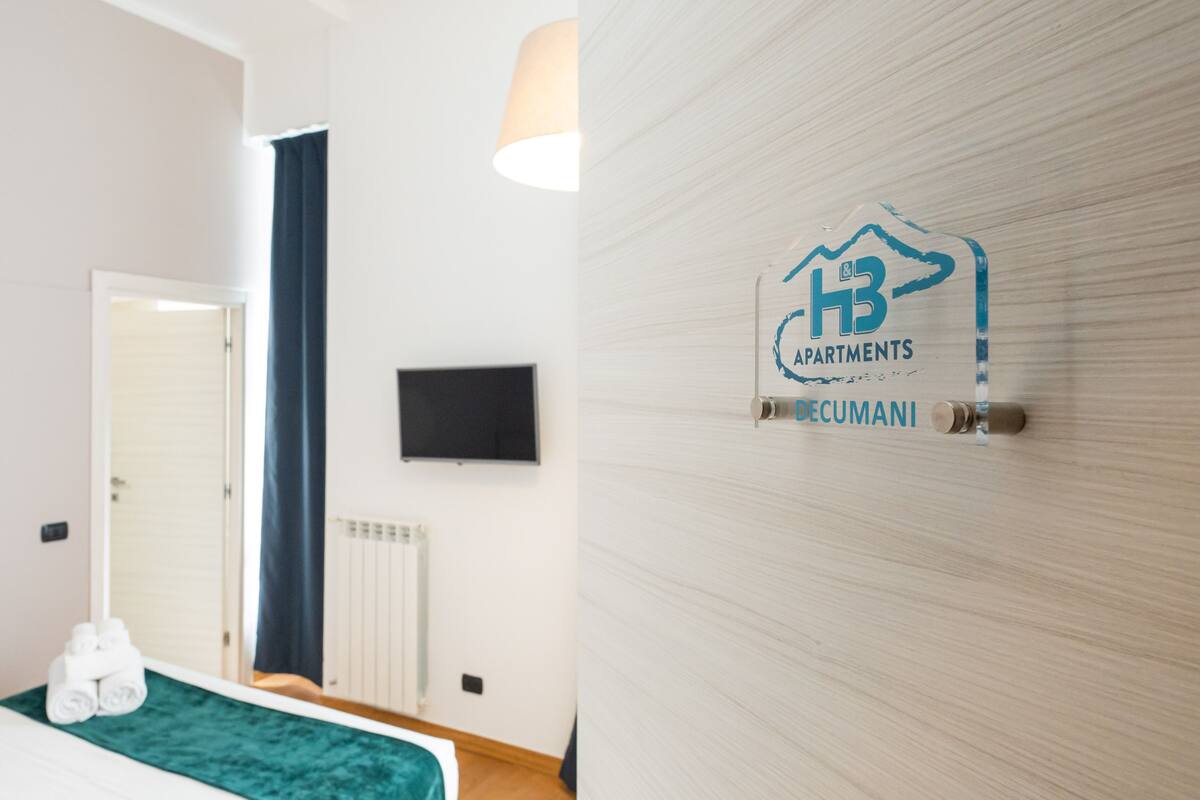 A hotel room with a bed, a TV mounted on the wall, and a logo for 'H3 Apartments Decumani'.