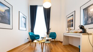 A modern room with a wooden floor, two blue chairs, a small round table, a white desk, and framed pictures on the walls. - Duomo Hermes Relais (Naples)