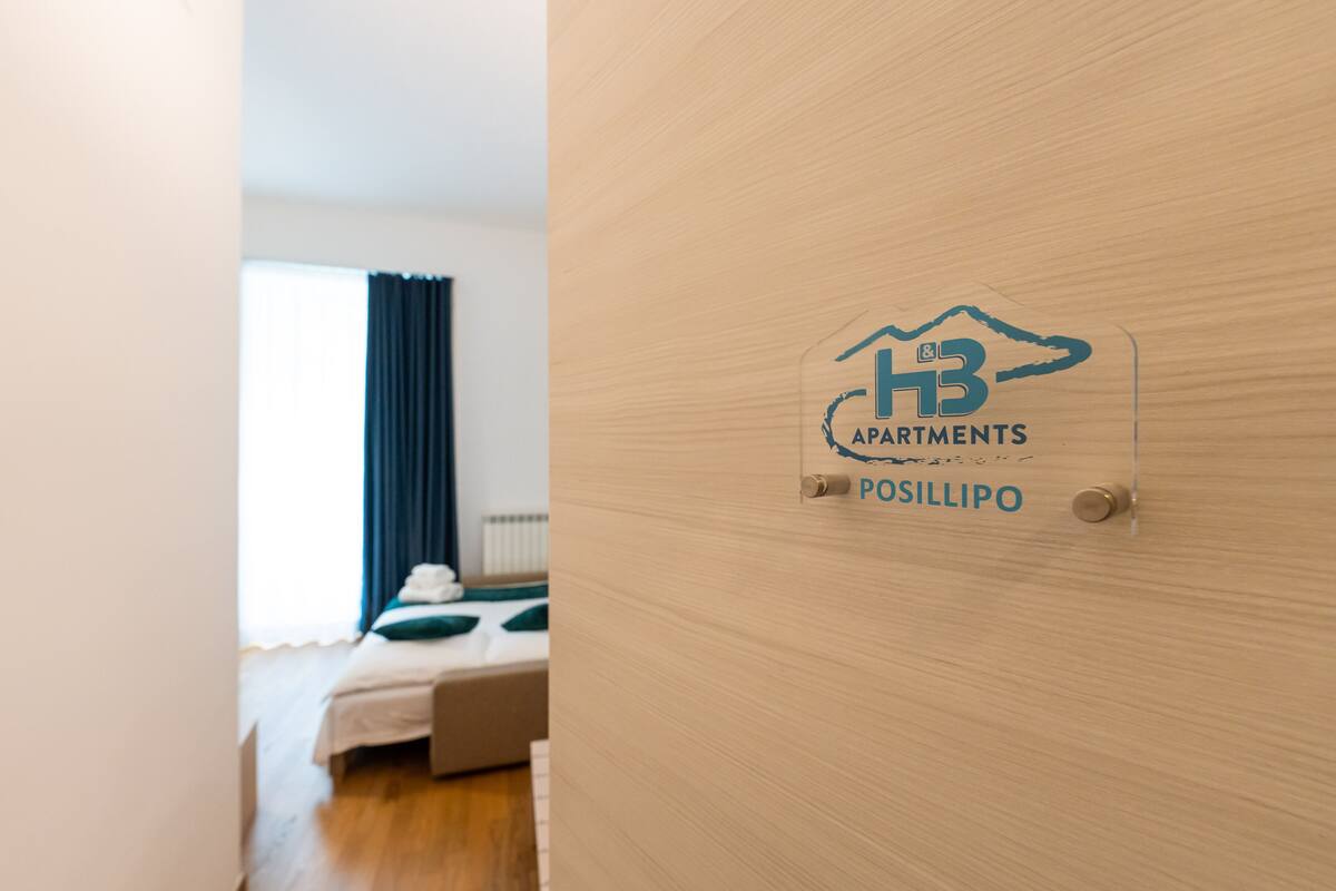 A hotel room with a bed, a window with curtains, and a wooden door with a sign reading "HB Apartments POSILLIPO".