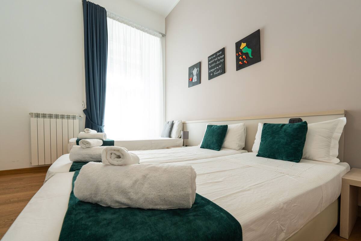 A hotel room with a large bed, two pillows, a bedside table, a window with curtains, and wall art.