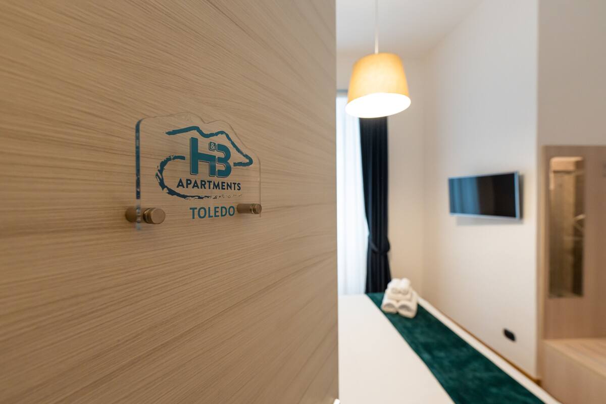 A hotel room entrance with a wooden door, a logo for "Toledo Apartments," and a TV mounted on the wall.