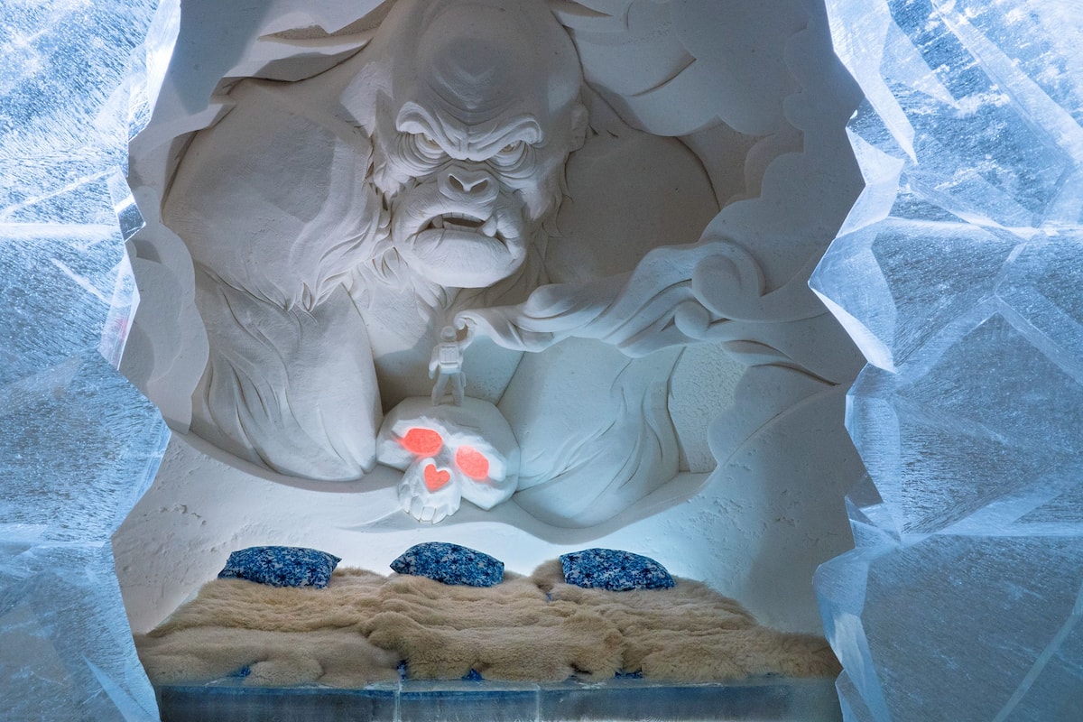 Une sculpture de glace représentant une figure barbu, une petite figure en train d’escalader, et trois coussins bleus posés sur une surface texturée.