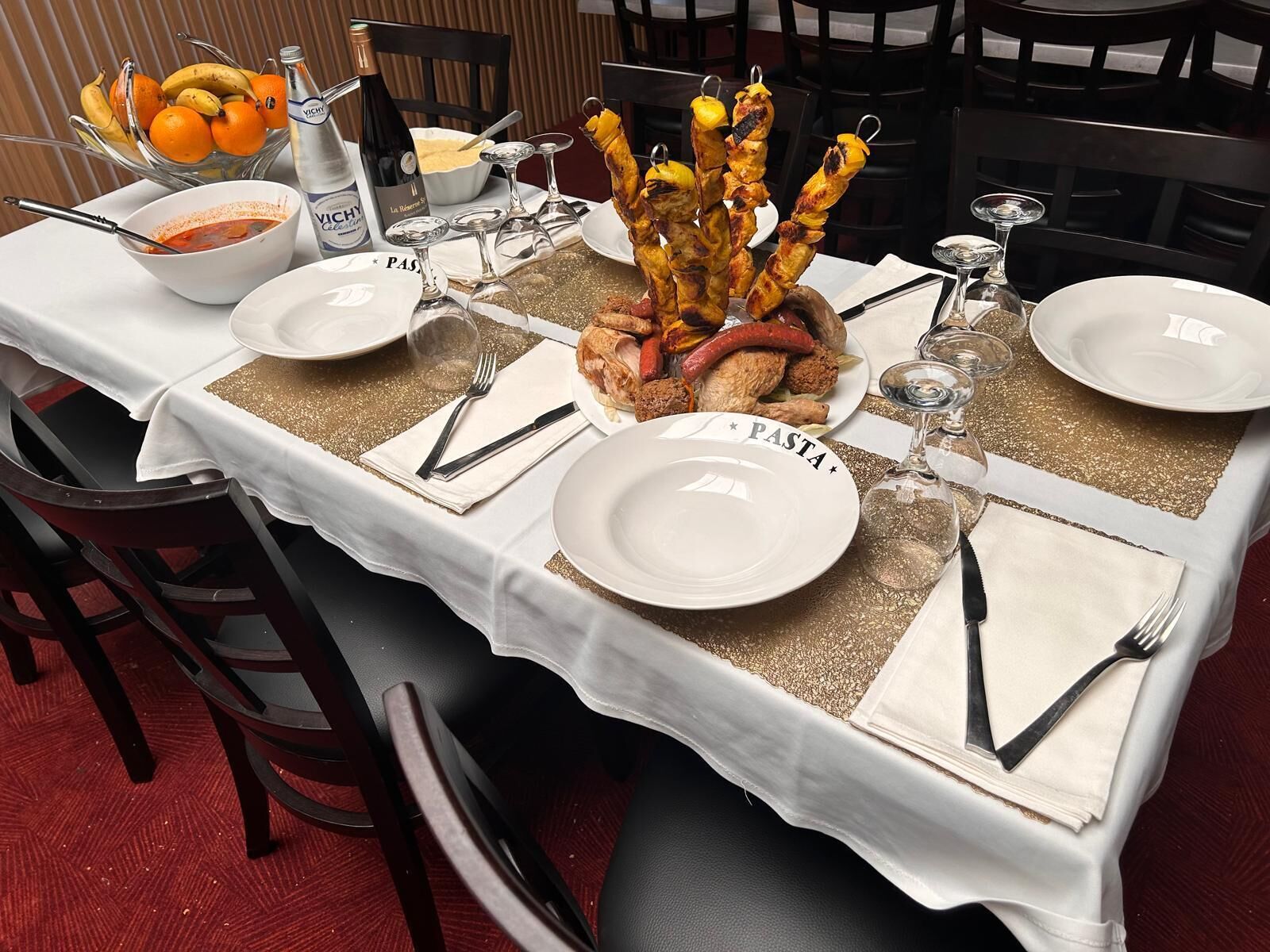 Una mesa de comedor dispuesta con platos, cubiertos y diversos alimentos, incluyendo brochetas y un bol de fruta.
