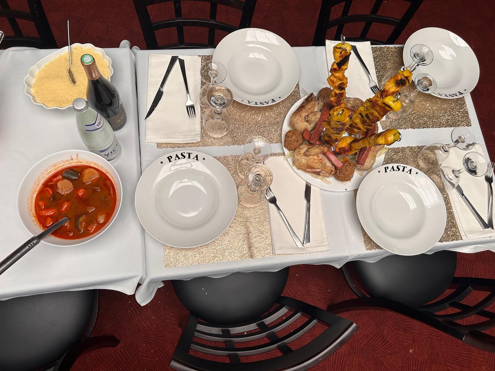 Une table dressée pour un repas, avec des assiettes étiquetées « pâtes », un bol de soupe et des brochettes de viande.