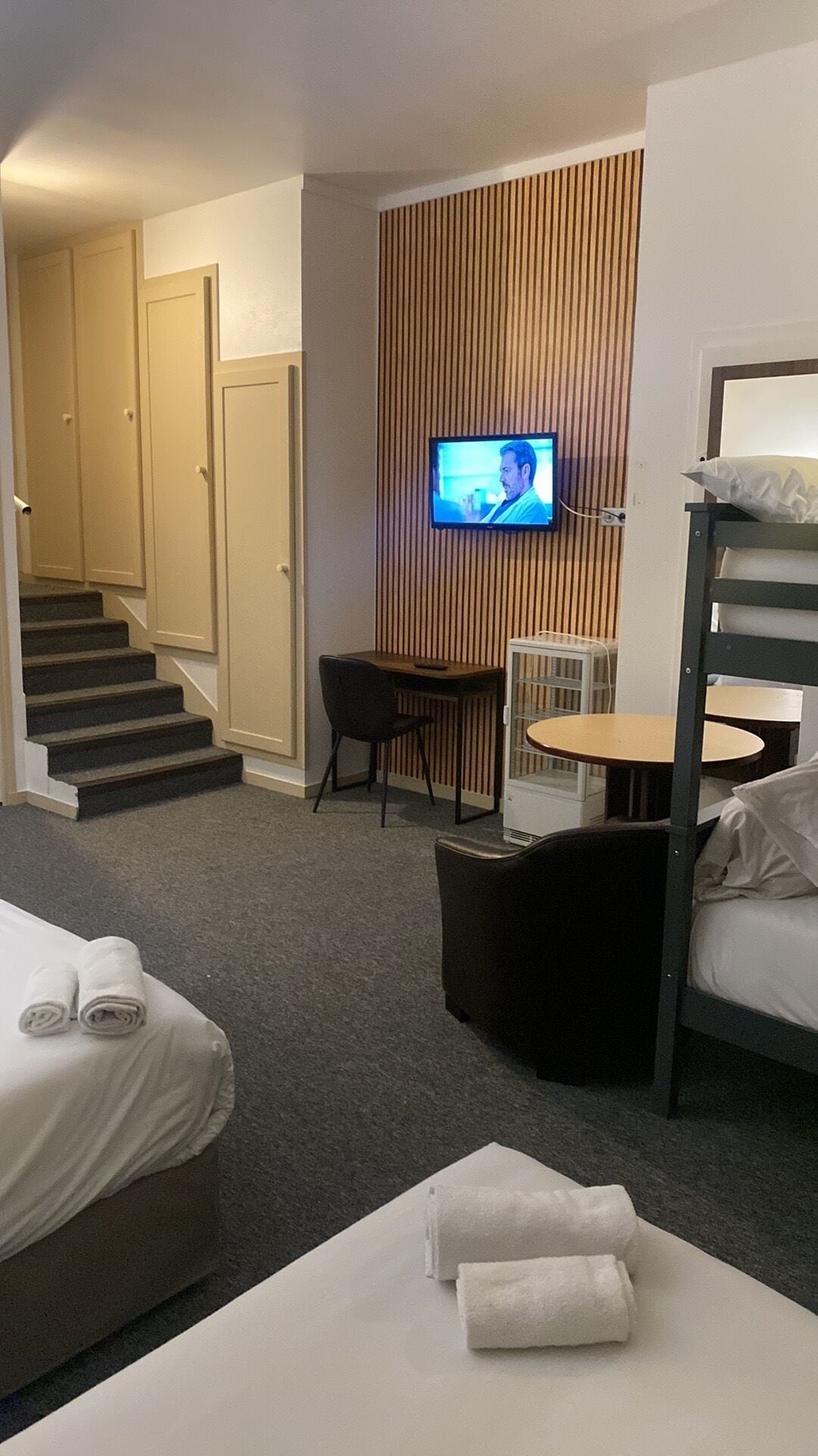A hotel room with a bed, a desk, a chair, a television mounted on a wooden panel, and a staircase leading to another room.