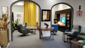 Een moderne woonkamer met een bank, fauteuils, een salontafel en een televisie. Er is een geel gordijn, een beeld en een plant.