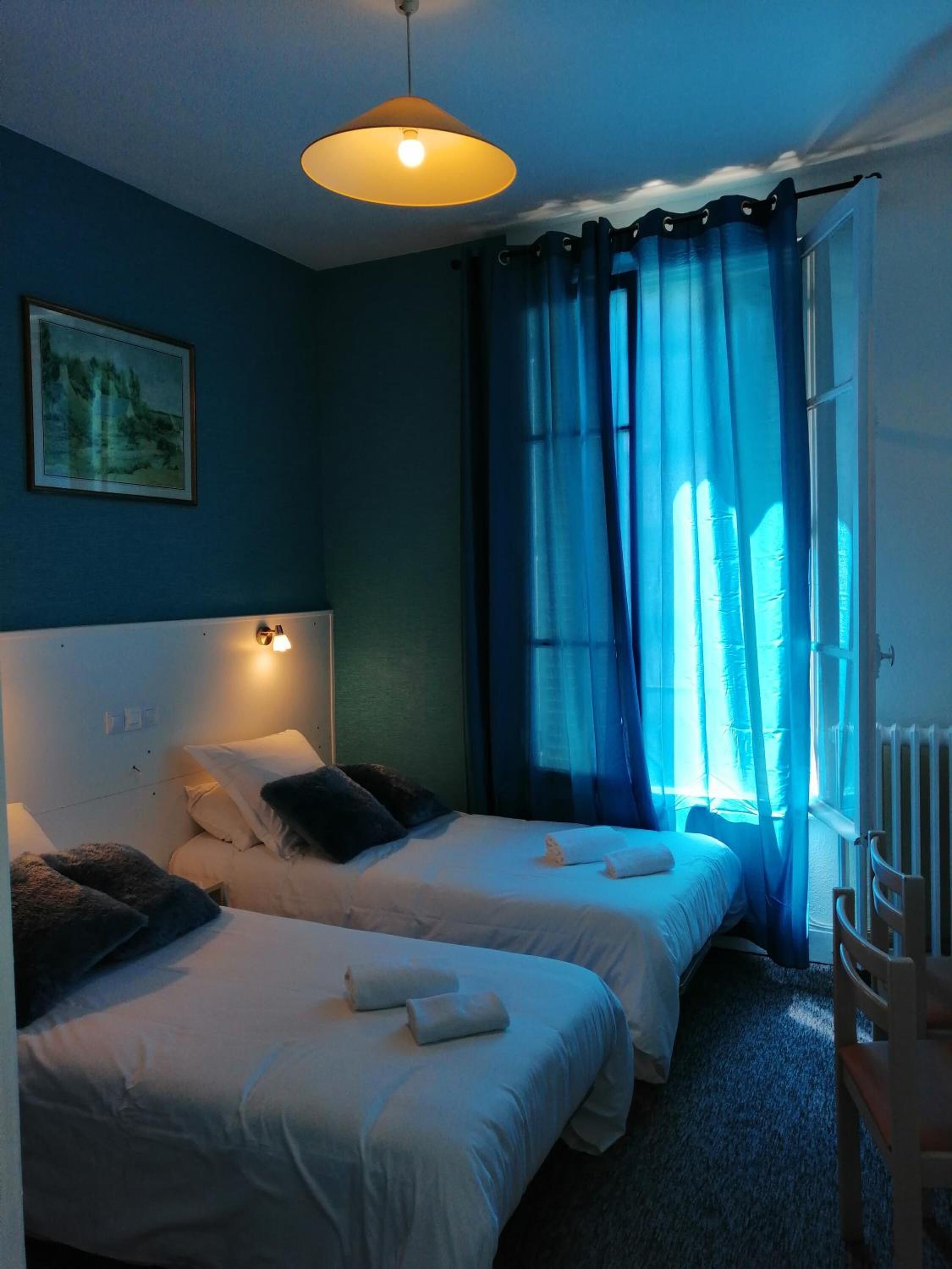 A bedroom with two beds, a painting on the wall, and a window with blue curtains.