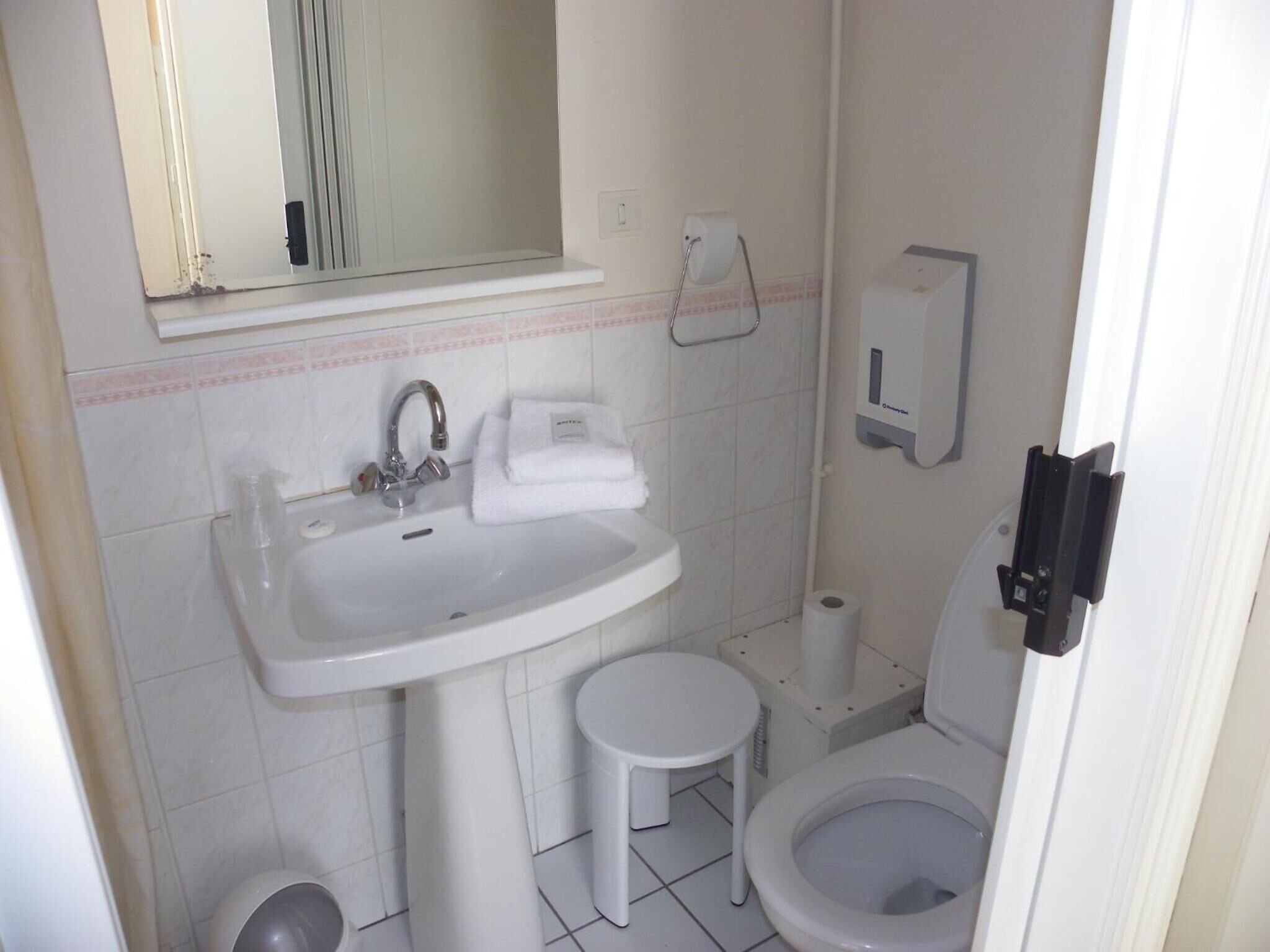 A small bathroom with a sink, mirror, towel, toilet, and a soap dispenser.