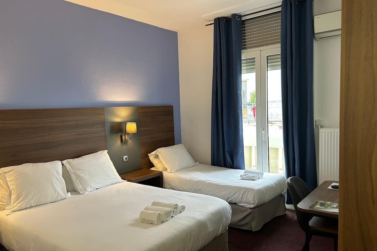 Hotelkamer met twee bedden, een nachtkastje met lamp, een kleine tafel met stoel, en uitzicht op de omgeving door een raam met blauwe gordijnen.