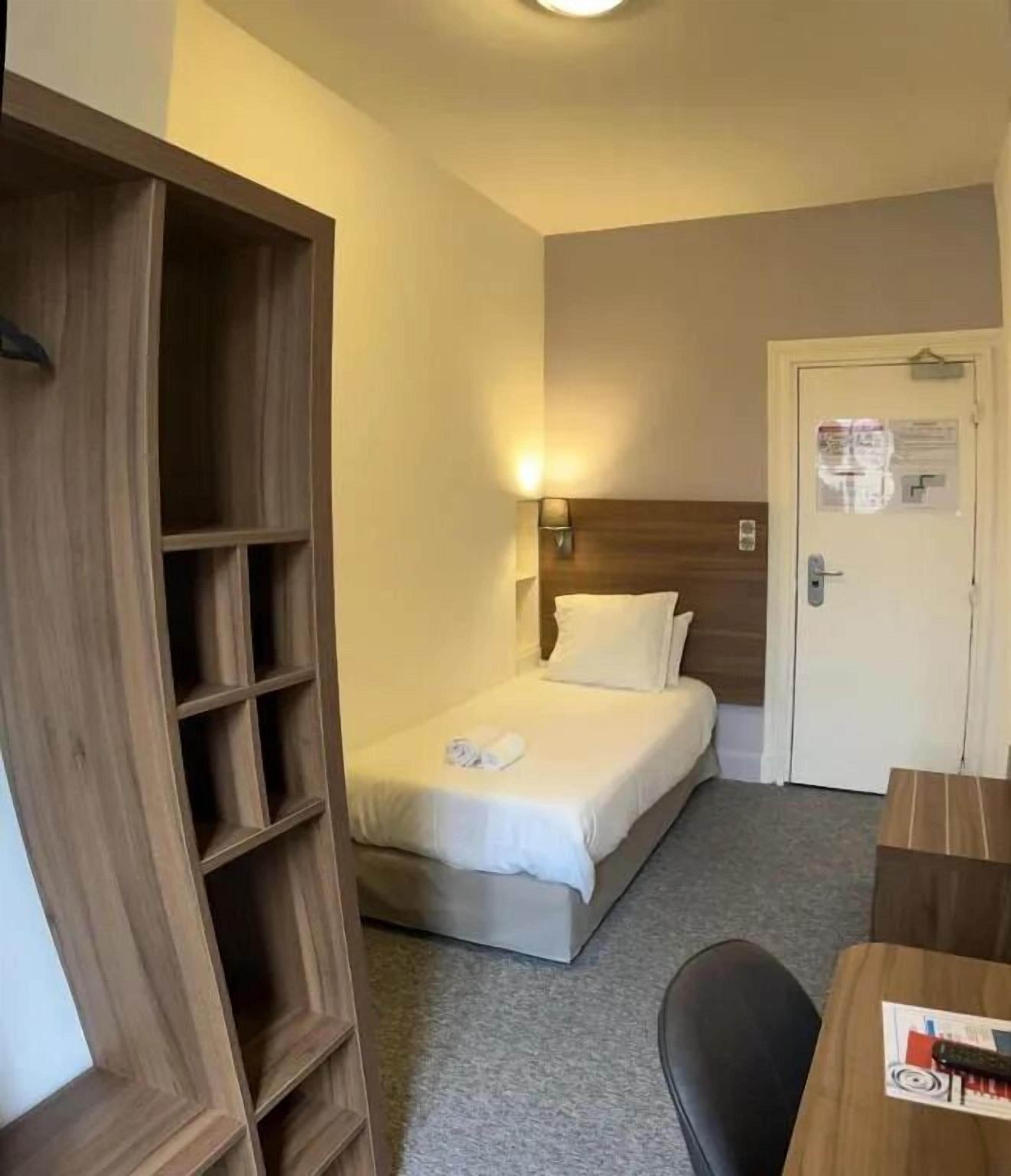 A small hotel room with a single bed, a wooden wardrobe, a desk with a chair, and a door with a notice.
