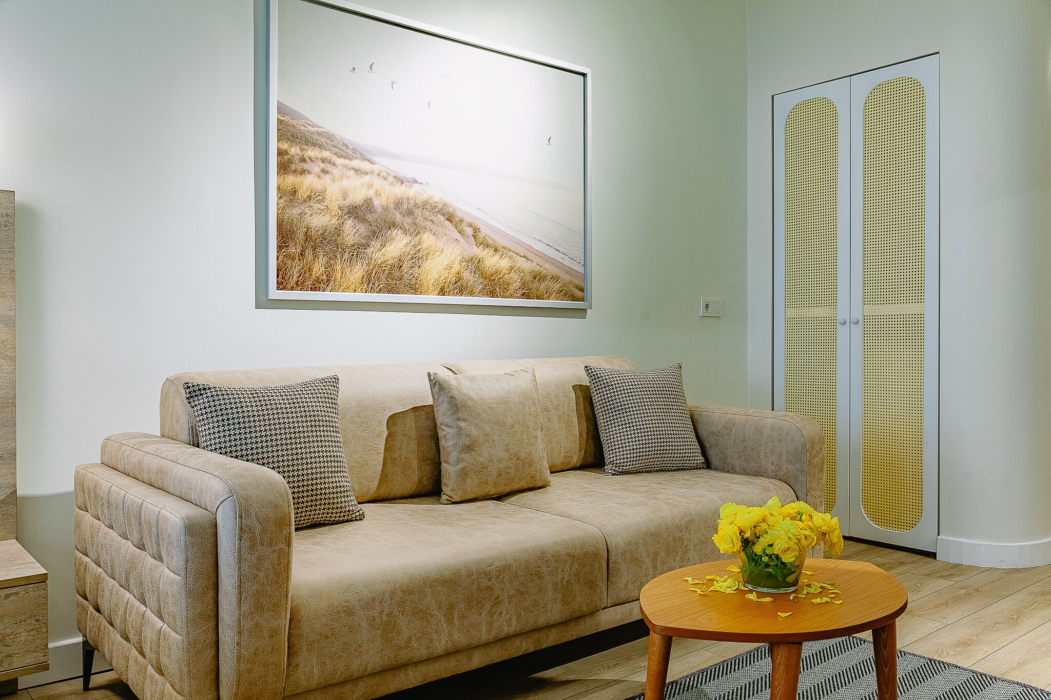 Un salon avec un canapé beige, un tableau représentant un paysage encadré, une table basse en bois ornée de fleurs jaunes et une porte au motif ajouré.