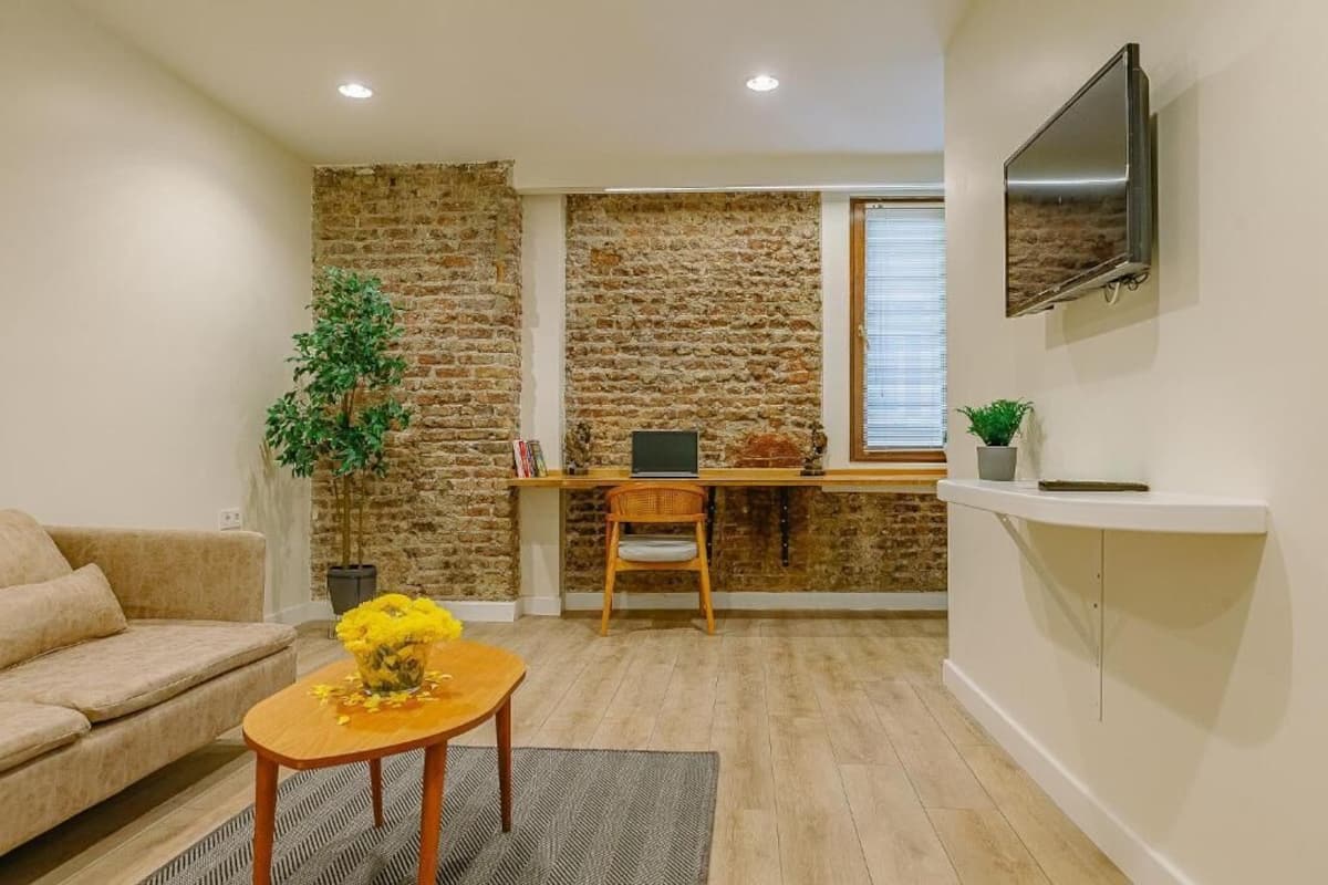 Un salon avec un canapé, une table basse ornée de fleurs, un bureau avec un ordinateur, un téléviseur fixé au mur et un mur en briques.