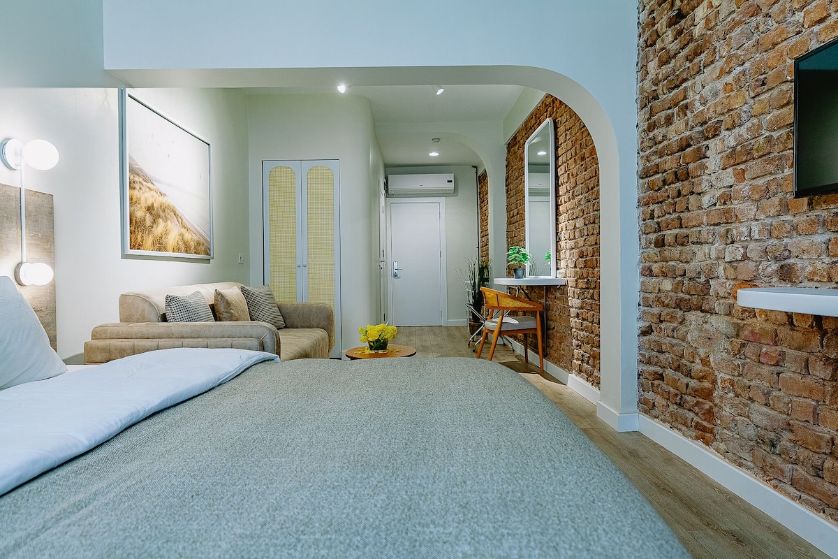 Une chambre moderne avec un grand lit, un coin salon avec un canapé, une petite table avec un vase et un mur en briques.