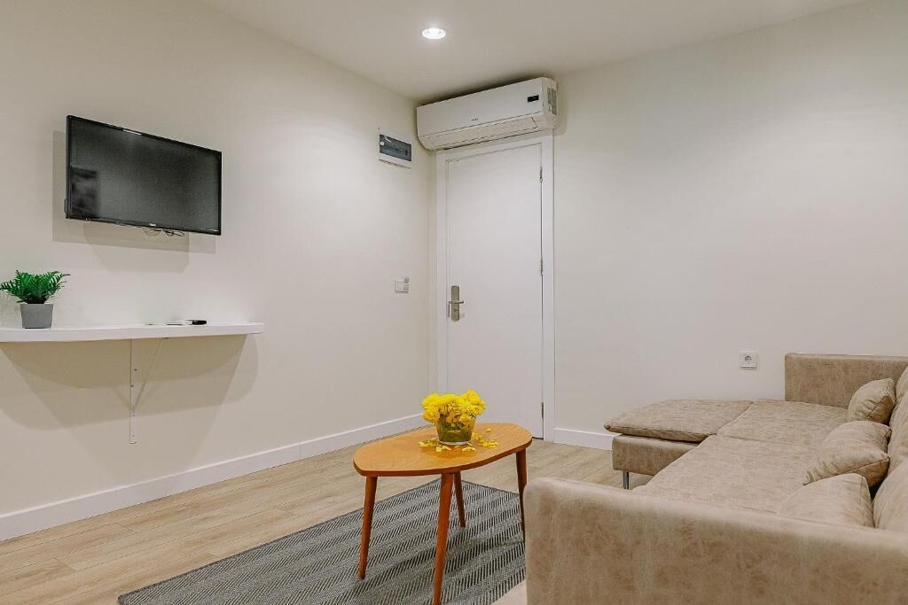 A modern living room with a flat-screen TV, a small wooden table with a vase of flowers, a beige sofa, and an air conditioning unit mounted on the wall.