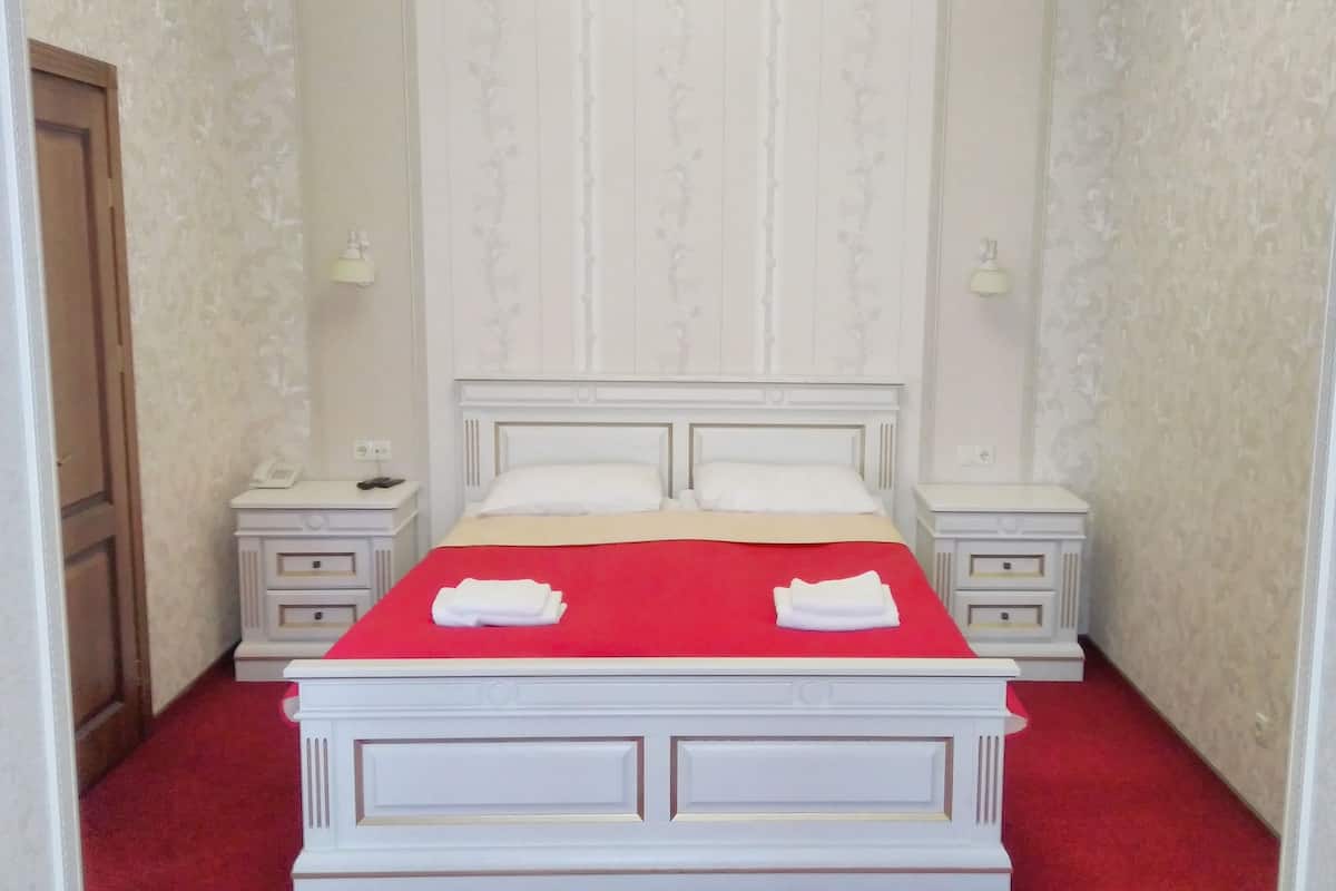 A neatly made bed with a red bedspread and two folded white towels, flanked by two bedside tables with lamps, in a room with patterned wallpaper and a wooden door.