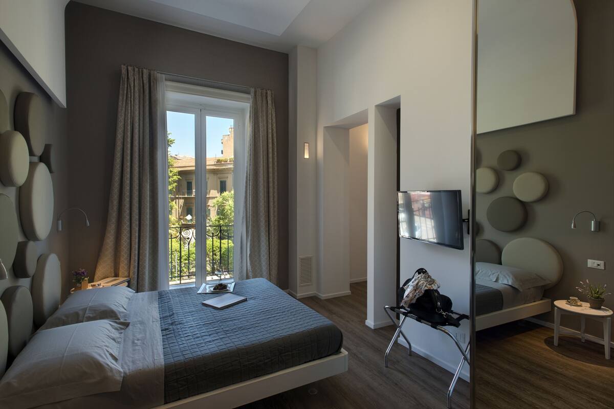 Una habitación de hotel moderna con cama, escritorio, silla, televisión y balcón con vistas a edificios.
