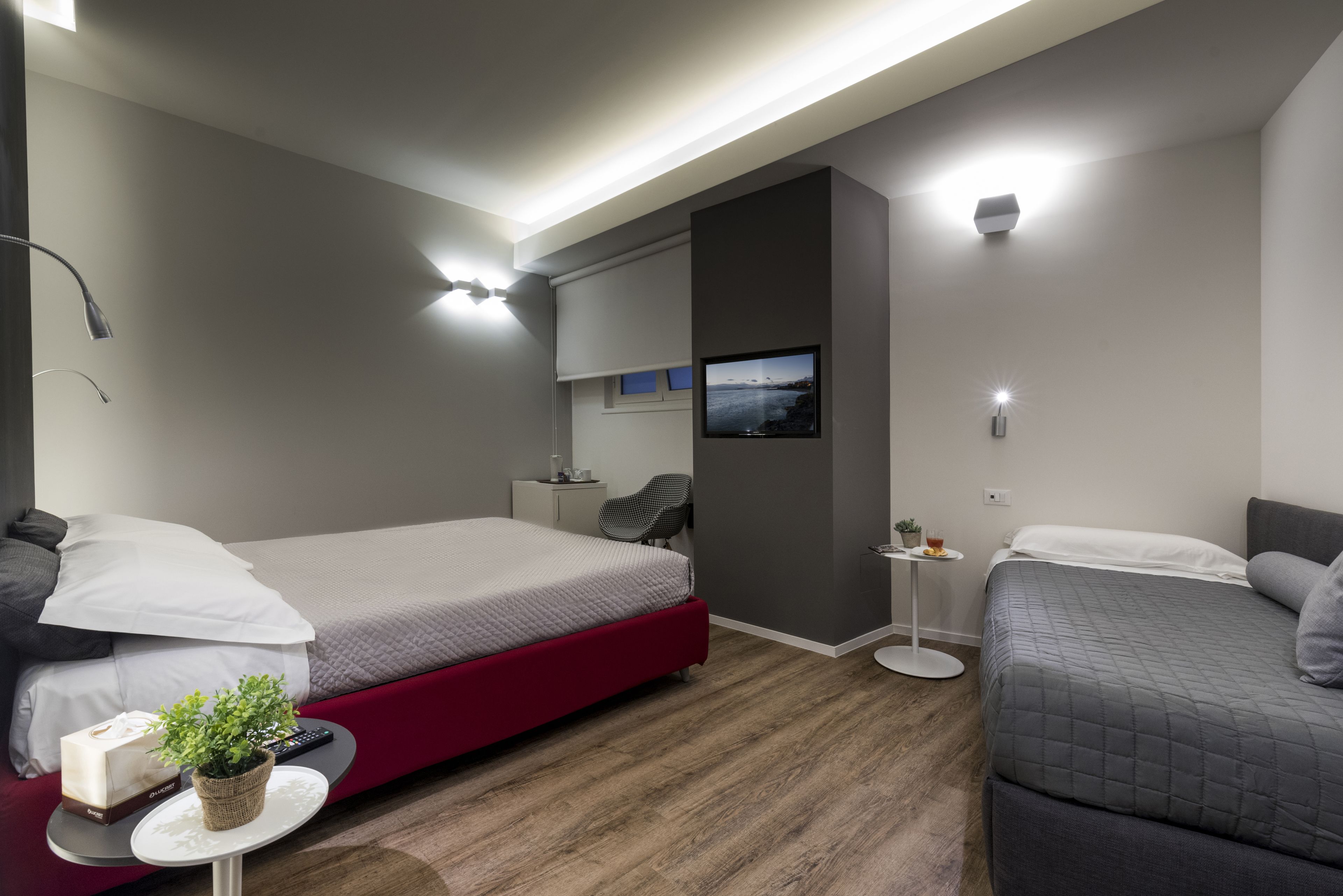 A modern hotel room with a large bed, a smaller bed, a TV mounted on the wall, and a small table with a plant.