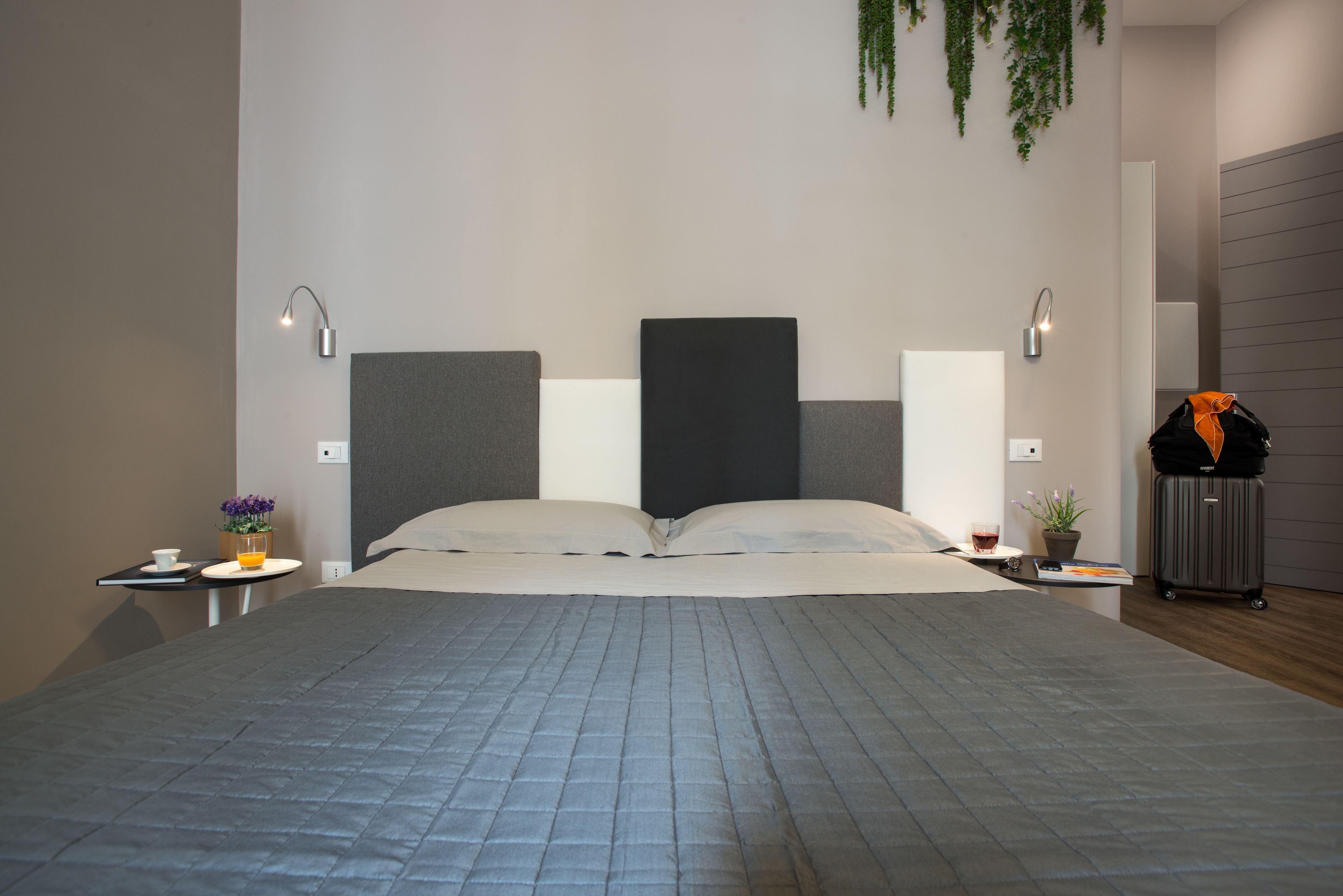 Ein modernes Hotelzimmer mit einem großen Bett, zwei Nachttischen, einem kleinen Tisch mit Tasse und Blumen, einer Topfpflanze und einem Koffer.