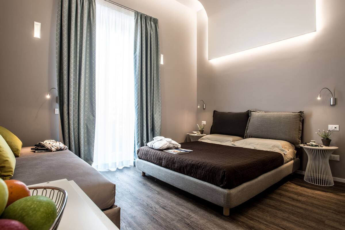 Una habitación de hotel moderna con una cama grande, una mesita de noche con lámpara, una pequeña mesa auxiliar con una planta y una cesta de frutas sobre una mesa.