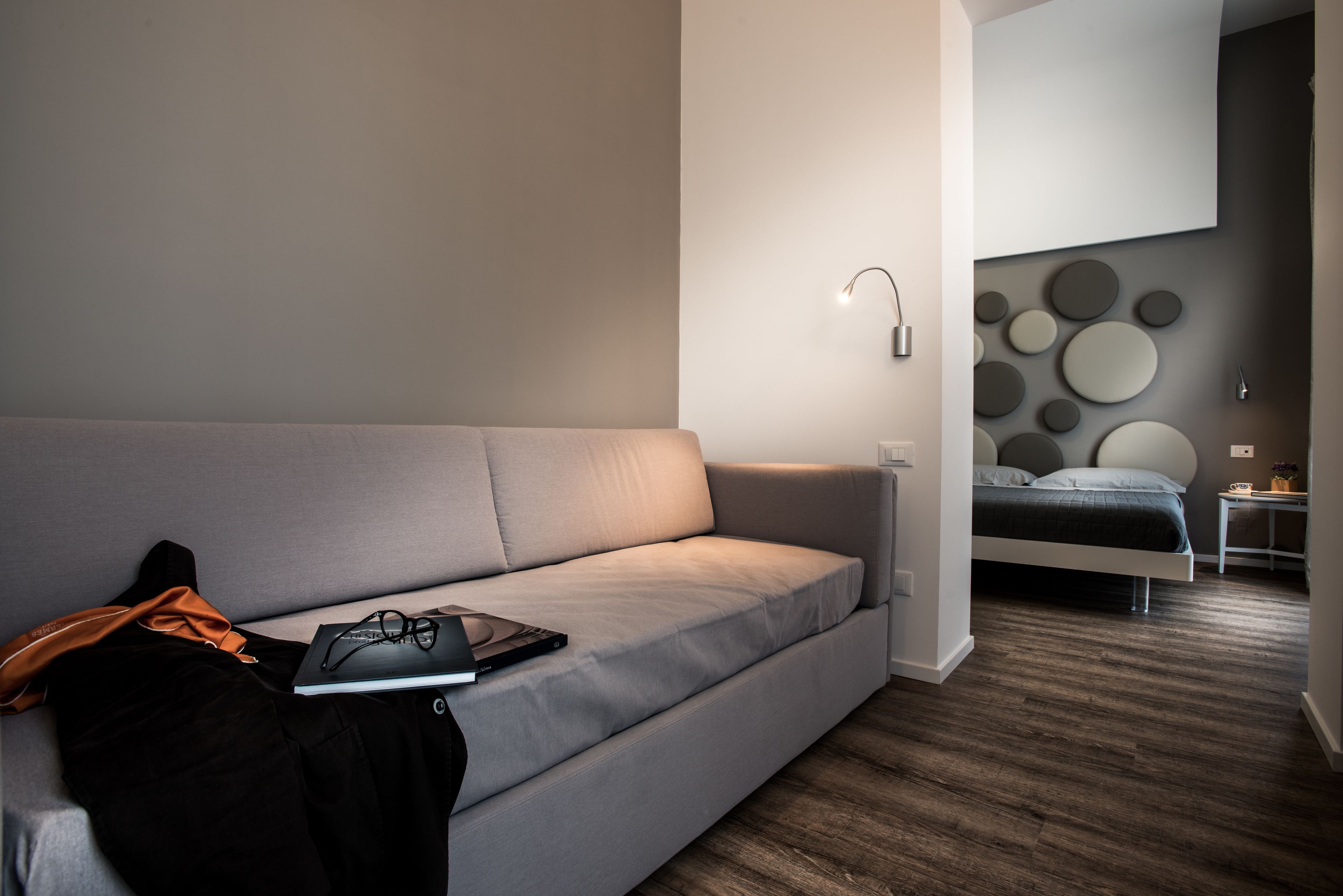 A modern, minimalist bedroom with a grey sofa, a small bedside table, and a bed with a grey headboard.