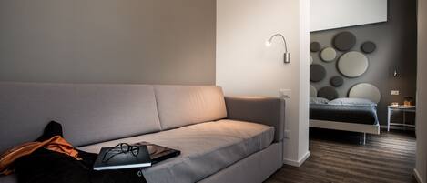 A modern, minimalist bedroom with a grey sofa, a small bedside table, and a bed with a grey headboard.