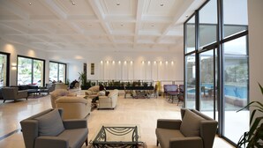 Een ruime lobby met een hoge plafond, grote ramen en diverse zithoekjes, waaronder banken en stoelen.