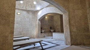 Een kamer met stenen muren, een hoog plafond en een centrale gewelfde deuropening, met een betegelde vloer. Er staan banken langs de muren en in de achtergrond een grote stenen wasbak.