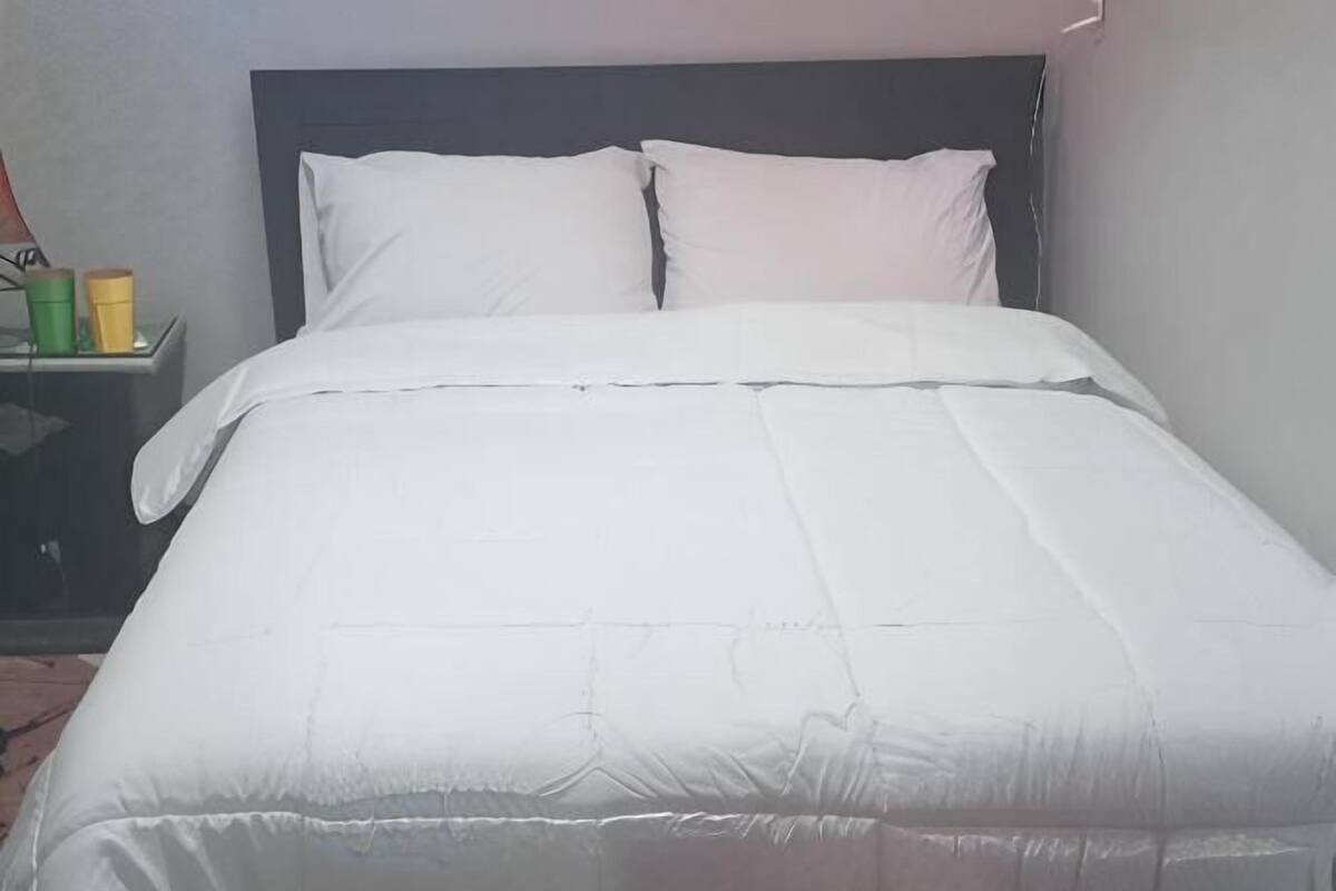 Una cama bien hecha con ropa de cama blanca en una habitación con una pared lisa y una mesita de noche pequeña con una lámpara.