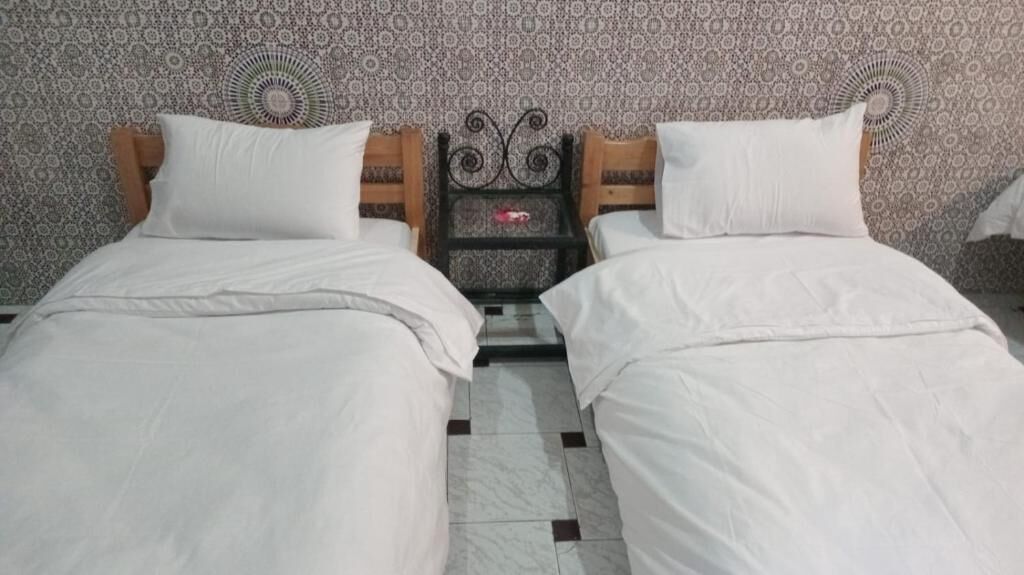 A room with two single beds, a small bedside table with a lamp, patterned wallpaper, and a tiled floor.